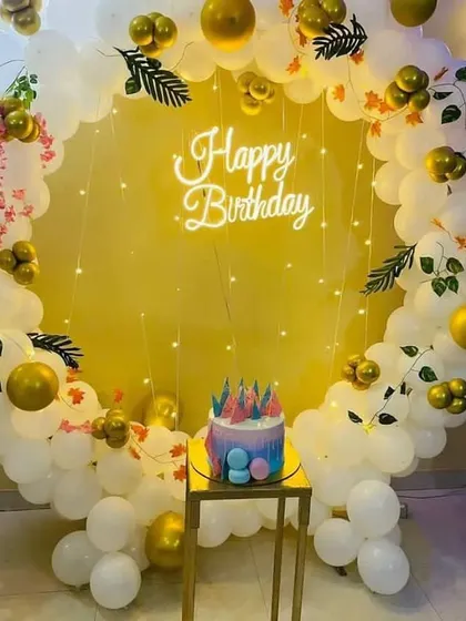 An elegant and classic birthday backdrop with a full ring of white and gold balloons, decorated with small leaves and fairy lights for a sophisticated look.