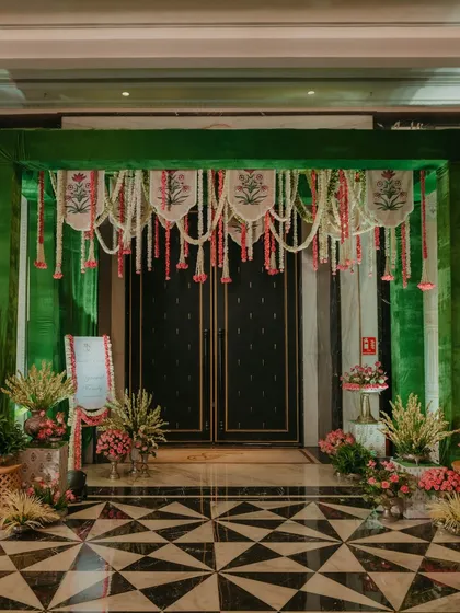 The entrance to the Myra ceremony, decorated with hanging floral strings and traditional block-printed banners, set against a rich green backdrop.