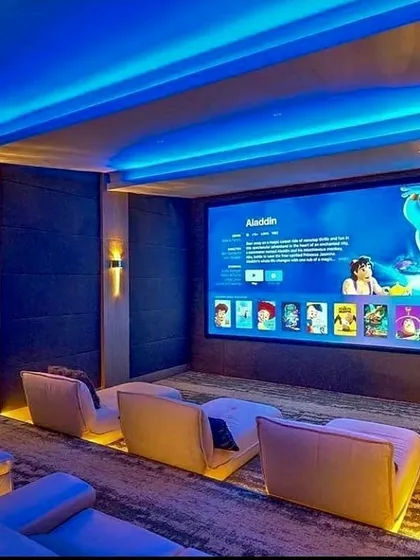 This contemporary media room is designed for both family enjoyment and critical viewing, featuring modern lounge seating with integrated floor lighting. The large-format screen is complemented by blue ambient cove lighting, creating an inviting atmosphere while ensuring excellent picture quality for everything from animated films to blockbuster movies.