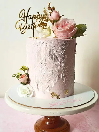 A tall, elegant pink cake with a modern, textured pattern. The cake is beautifully decorated with fresh flowers and a touch of gold leaf for a sophisticated finish.