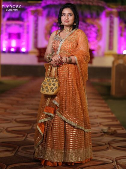 A full-length view of the shimmering orange lehenga. Paired with a matching potli bag, it's a complete and glamorous look for any festive occasion.