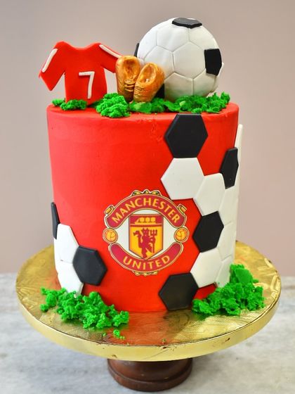 A cake for a true football fan. This design is dedicated to Manchester United, featuring the team's logo, a jersey, and soccer balls, all made from edible fondant.
