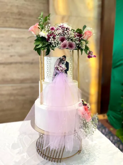 A modern and romantic three-tier wedding cake. This design features a custom photo of the couple, a delicate tulle veil, and a beautiful fresh flower arrangement on top, creating a personalized and elegant centerpiece.