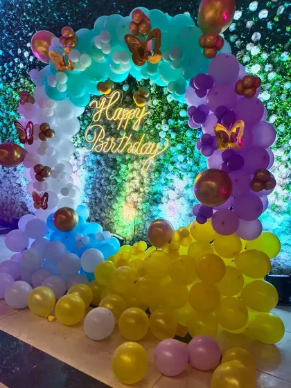 A beautiful butterfly-themed birthday setup against a green wall. The rainbow-colored balloon arch with butterfly foils creates a magical garden feel.
