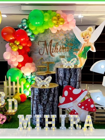 A whimsical fairy-themed third birthday party. The decor includes a Tinkerbell cutout, a forest backdrop, log-style plinths, and a large light-up number '3', creating an enchanted woodland scene.