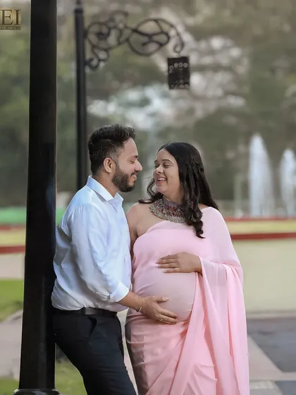 A sweet, shared look between the couple during their outdoor park photoshoot. The mom-to-be is elegant in a pink saree.