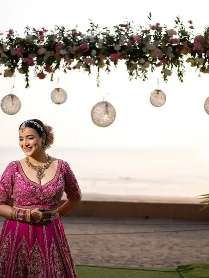 The bride stands smiling under her beautiful floral wedding arch, with the golden hour sun and the sea behind her.