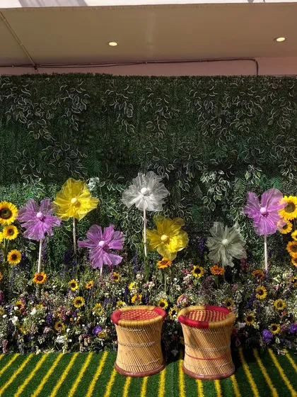 A beautiful photo wall made of a green foliage backdrop, decorated with sunflowers and large fabric flowers, complete with traditional 'mooda' seats.