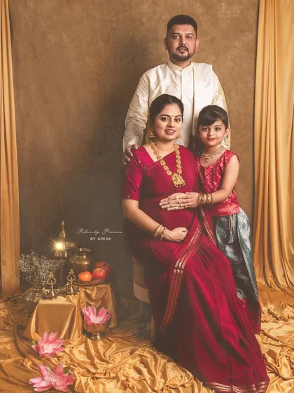 A regal family portrait celebrating a traditional Indian maternity journey. The mother, father, and daughter are dressed in elegant ethnic wear, surrounded by cultural props like brass vessels and lotuses, creating a timeless memory.