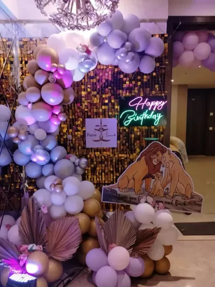 A Lion King themed party entrance with a gold shimmer wall, purple and gold balloons, and a "Happy Birthday" neon sign.
