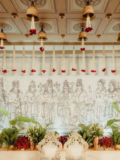 The serene backdrop of the mandap, featuring a large, hand-drawn panel depicting revered deities. Hanging temple bells and jasmine garlands frame the artwork, adding to the sacred and artistic ambiance of the ceremony space.