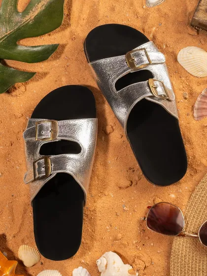 A beach-themed flat lay of silver double-buckle sandals on sand. The styling with seashells and sunglasses evokes a summer holiday feel.