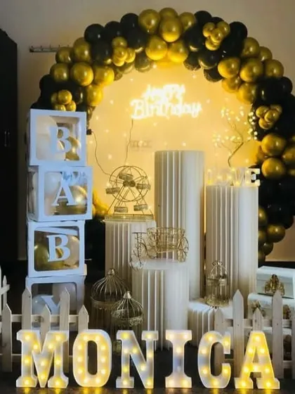 A chic black and gold birthday theme for Monica. The setup includes a balloon arch, 'BABY' blocks, a white picket fence, and various elegant props on display pedestals.