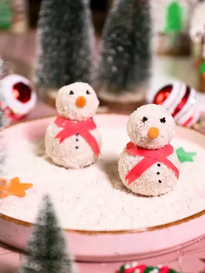 Cute snowman-shaped coconut laddoos sitting in a bed of "snow" on a pink plate. This playful and creative dessert shot is perfect for a fun-filled Christmas menu.