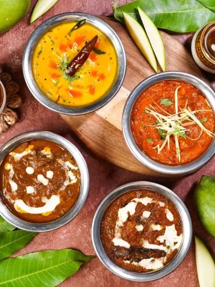 An assortment of mango-based Indian curries and dals, shot from above. The composition with raw mangoes and leaves highlights the seasonal theme of the menu.