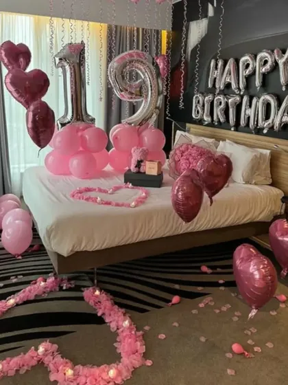We do birthday surprises too! This 19th birthday room decoration features pink and silver number balloons, heart-shaped foil balloons, and flower petal hearts on the bed.