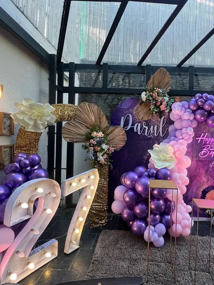 A chic and trendy decoration for a 27th birthday, using a rich purple marble-effect backdrop, metallic balloons, and dried palm leaf accents.