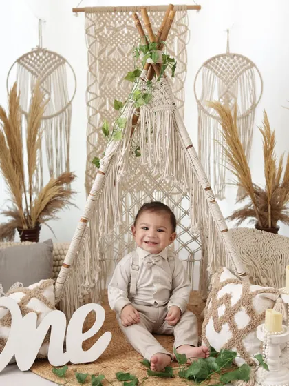Officially one! A proud and happy one-year-old boy sits in front of a large "one" sign, surrounded by a stylish boho setup with macrame and pampas grass.