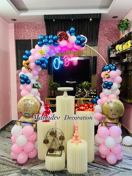 An "Oh Baby" gender reveal party setup in a living room. The decoration features a pink and blue balloon arch, 'boy' and 'girl' baby foil balloons, and a neon sign, making it perfect for the big reveal.