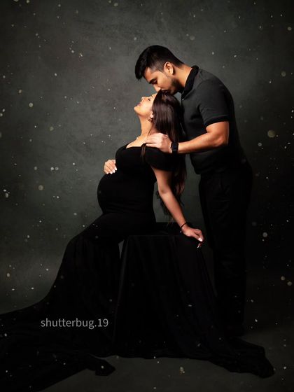 A classic and romantic pose for an expecting couple. Dressed in black, the father-to-be kisses his partner's forehead as she looks up at him, creating a timeless memory against a starry-night-inspired backdrop.