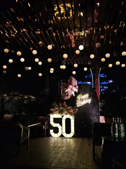 A wide shot of a 50th birthday party on a beautiful wooden deck. The string lights on the ceiling and the glowing decor create a magical and inviting atmosphere.