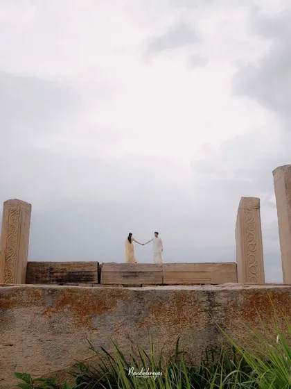 A wide shot of a couple holding hands atop a stone wall, framed by ancient pillars and an expansive sky.
