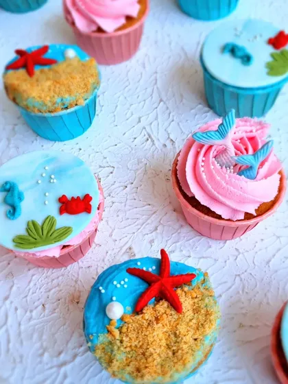 A variety of underwater-themed cupcakes. These show different topping styles, from buttercream swirls with fondant tails to smooth fondant tops with beach scenes.