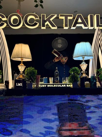 A bartender expertly pours a drink at a stylish black and white themed bar. The setup, complete with classic lamps and Art Deco pillars, provides a sophisticated backdrop for our craft.