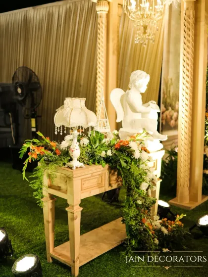 A close-up of a vintage-inspired decor corner. A classic table lamp, a cherub statue, and a cascade of greenery and flowers create a small moment of timeless beauty.