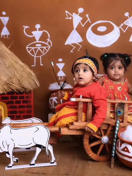 A playful and creative Janmashtami themed shoot. Two babies dressed as little Krishnas sit in a miniature wooden bullock cart, surrounded by a rustic village backdrop, showing the fun and imaginative sets we create.