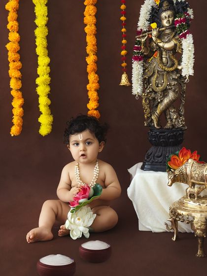 A baby dressed as Krishna holds a lotus flower, looking inquisitively at the camera in a clean, simple setup.