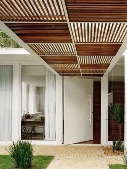 A stunning wooden rafter pergola creates a covered walkway leading to the entrance of a modern home. The checkered pattern of the rafters adds architectural interest and provides partial shade for the path.