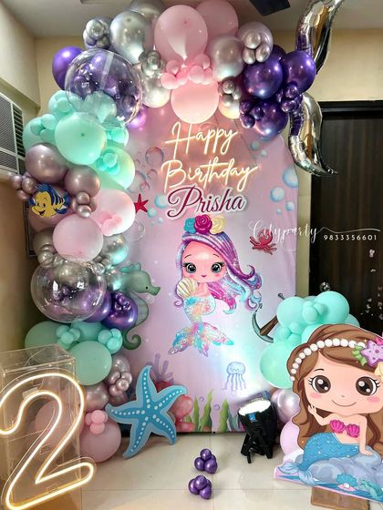 A cute mermaid theme for a 2nd birthday party. The decor features a custom backdrop with a cartoon mermaid, a balloon arch in sea-inspired colors, a light-up marquee number '2', and a mermaid character standee.