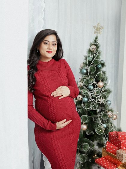 A festive solo maternity portrait. This mom-to-be looks beautiful in her red sweater dress, standing beside a Christmas tree, celebrating this special time of year.