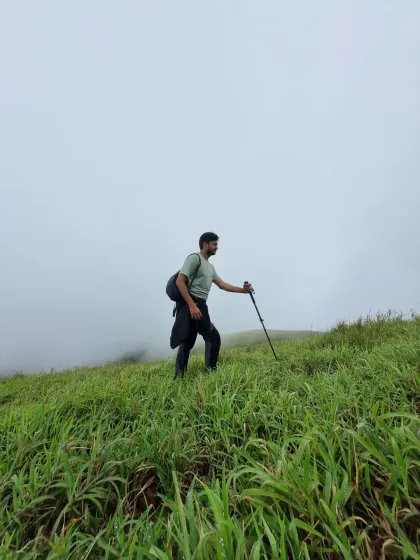 A trekker pauses on a grassy slope on the way to Nethravathi Peak, trekking pole in hand. The thick fog behind him shows the challenging yet beautiful weather conditions we often encounter.