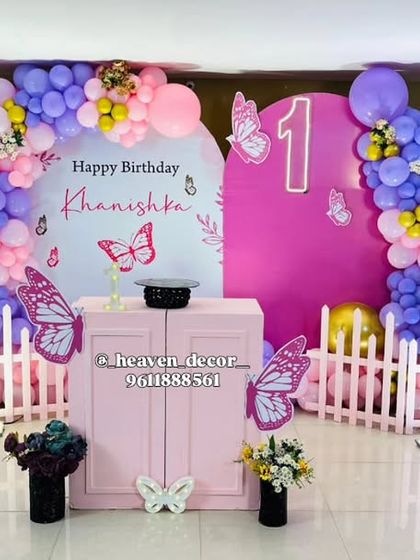 A lovely butterfly garden setup for a first birthday, featuring large pink wing cutouts and a matching picket fence. The backdrop is personalized, and the scene is completed with a colorful balloon arrangement.