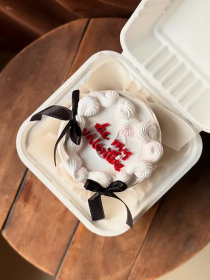 Celebrate your love with a romantic Valentine's Day bento cake. This design features "Be my Valentine" written in red on a white cake, decorated with piped frosting and elegant black ribbons, all nestled in its own lunchbox.