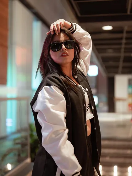 This portrait captures a cool, confident attitude. The model's varsity jacket and sunglasses, combined with the blurred urban background, create a classic street style look.
