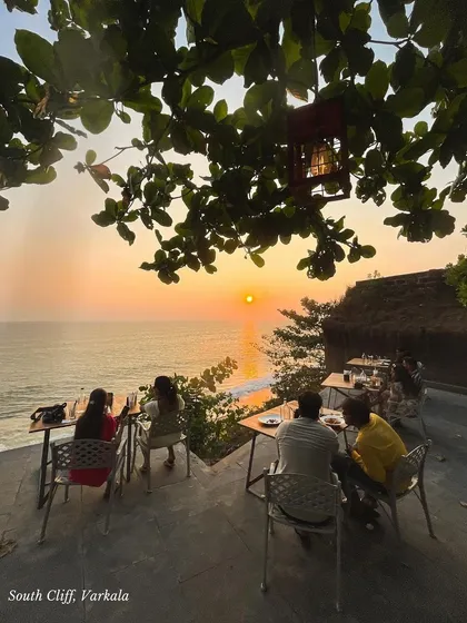 A group dines at a cafe on the South Cliff of Varkala as the sun sets over the ocean. It's a perfect blend of good food, great company, and breathtaking views.