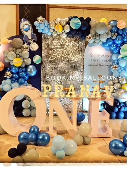 We crafted an out-of-this-world space adventure for Pranav's first birthday. The setup features a sparkling silver backdrop, balloon garlands in cosmic blues and greys, and custom props like a giant moon, astronaut cutout, and rocket ship to create a stellar atmosphere.
