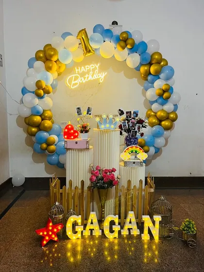 A beautiful ring backdrop for a first birthday celebration. The balloon ring in blue, white, and gold chrome frames a display of photos and props on plinths, with a 'Happy Birthday' neon sign and light-up name.
