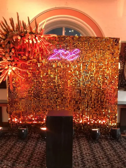 We design eye-catching photo booths that become a highlight of any event. This setup features a shimmering golden sequin wall, a custom neon logo, and dramatic floral arrangements, providing a perfect spot for guests to capture memories.