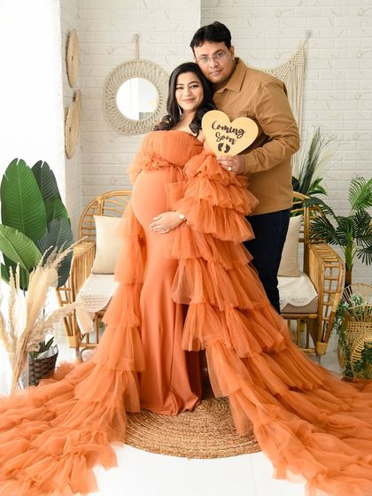 A sweet couple's portrait in our boho-themed studio. They are holding a "Coming Soon" sign, and the mom-to-be's orange ruffled gown adds a vibrant touch to the earthy decor.