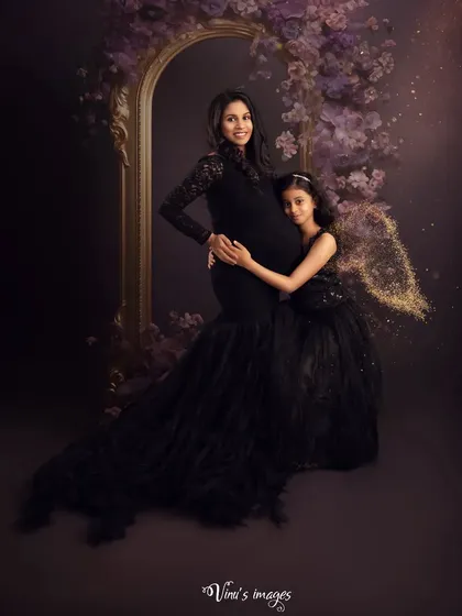 A dramatic and magical maternity portrait. The mother and daughter are dressed in elegant black gowns, with the daughter's fairy wings adding a touch of enchantment to this studio session.