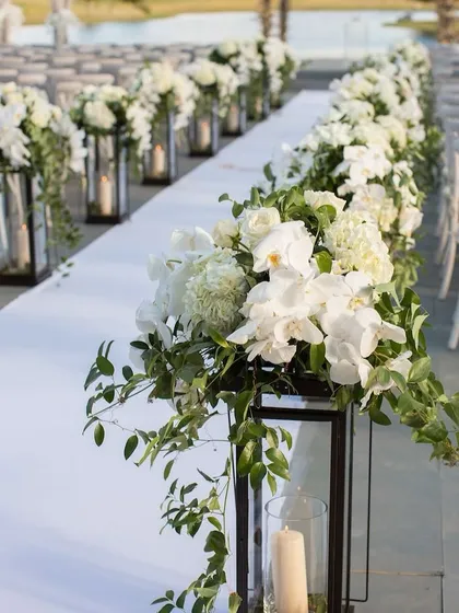 A close-up of an elegant white wedding aisle. The path is lined with beautiful arrangements of white flowers and greenery in modern black lanterns, creating a sophisticated and timeless look.