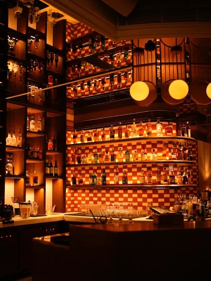 The bar at night, fully stocked and glowing. The checkered tile and warm lighting create a classic, inviting backdrop for an evening of great drinks.