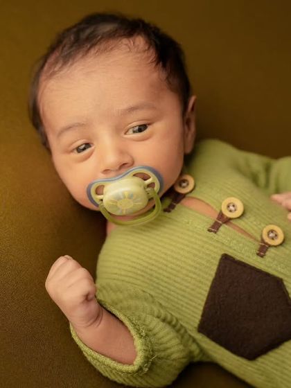 An awake newborn in a green knit outfit with a pacifier, looking curiously at the camera on an olive green background.