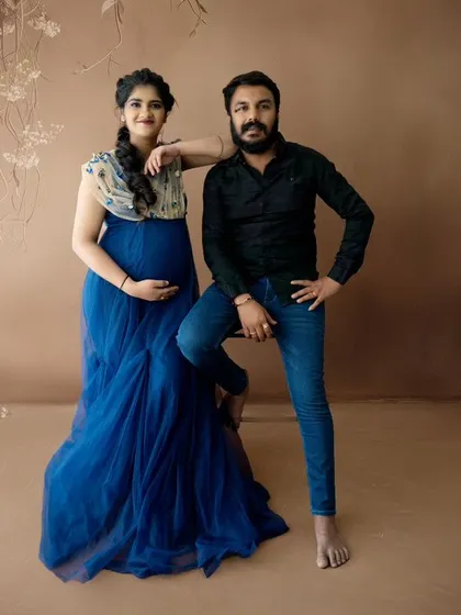 A stylish and confident couple's portrait against a simple brown backdrop, with the partner seated and the mom-to-be standing.