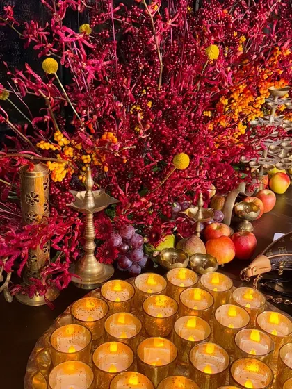 A close-up of a Diwali decor setup, with a tray of glowing candles, brass lamps, and vibrant red and yellow foliage.
