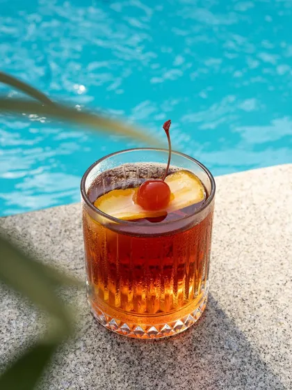 A dark, moody cocktail by the pool. The contrast between the deep amber drink and the bright blue water creates a sophisticated and relaxing summer image.